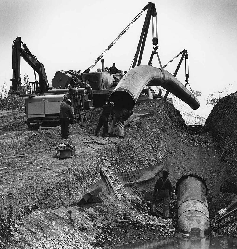 Old black and white photo of 1975 Ahtna Construction workers installing the first piece of the Trans-Alaska Pipeline System at Tonsina River