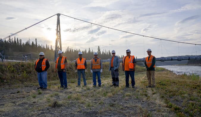 7 workers in hardhats and vests pose before Trans Alaska Pipeline.
