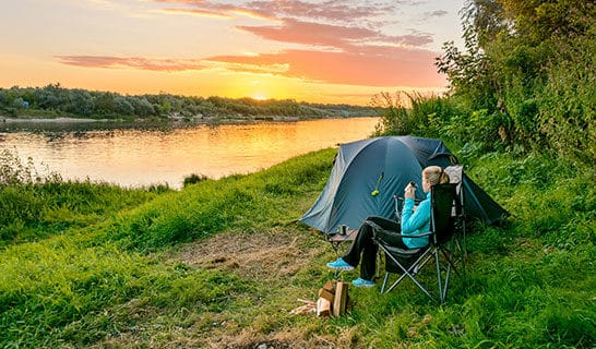 Woman sipping coffee next to her tent while enjoying the sunrise over a lake.