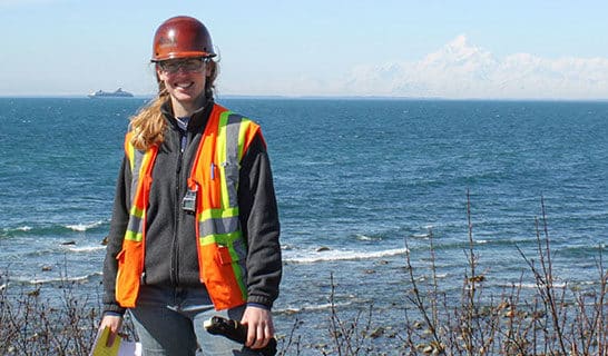 Female Ahtna worker in hardhat and safety vest in front of the water with Alaskan mountains behind.