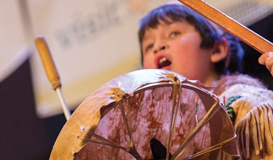 Young Alaska native boy beats a drum in ceremonial dress.