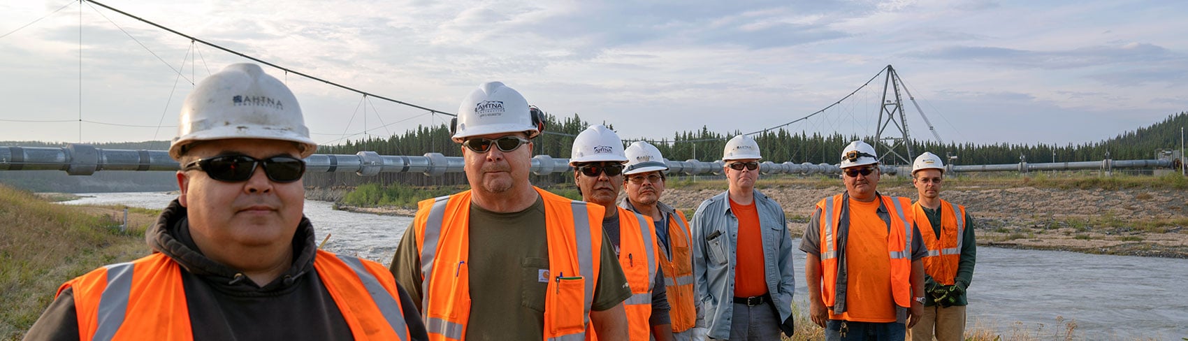 7 men in the Alaska wild working in Ahtna hardhats and safety vests. A river with the trans Alaska pipeline behind them.