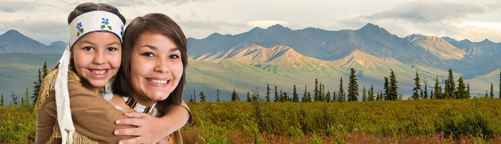 Woman with girl hugging her shoulders, both in traditional native dress, with Ahtna mountains in the background.