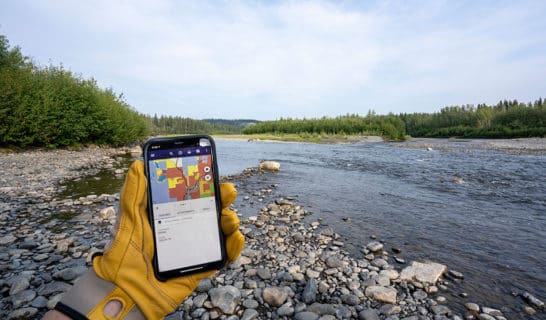 Ahtna's Regional Map App loaded on a phone held by a gloved hand in front of an Alaskan river.