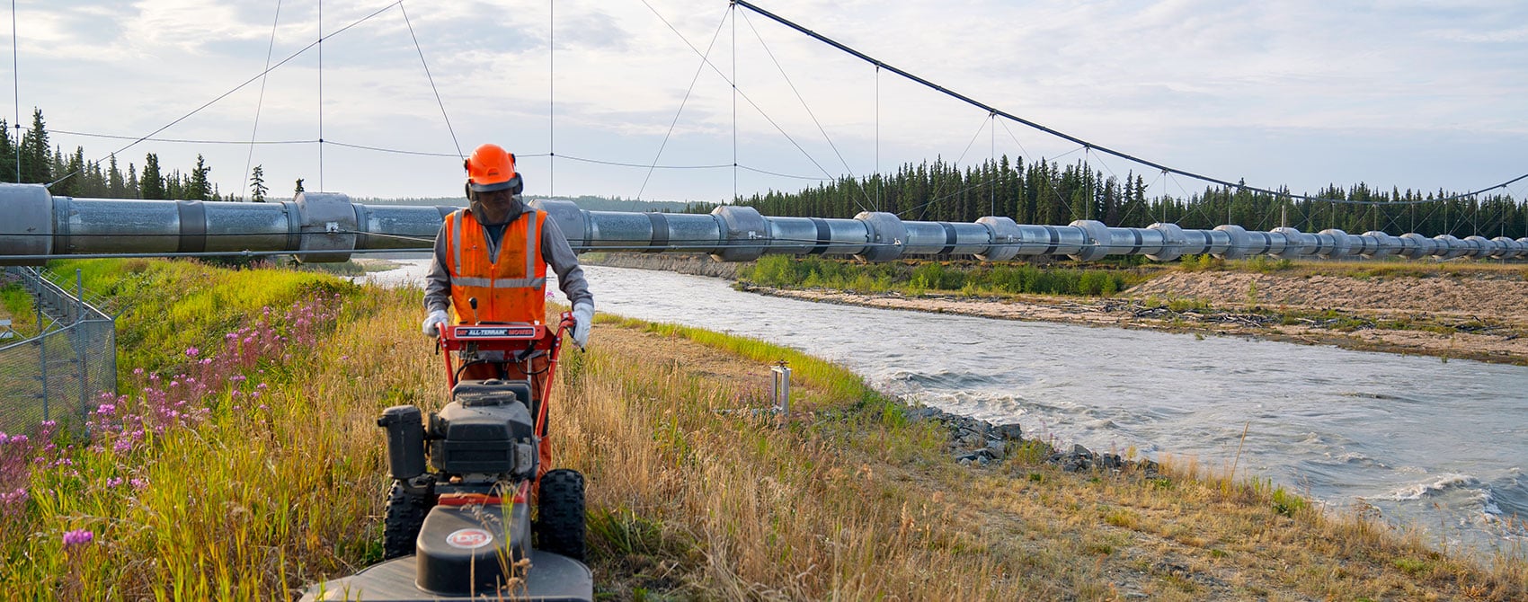Workers mows back grass along the river with the Alaska pipeline crossing behind.